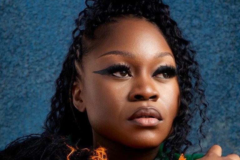 Close-up portrait of musician Sampa the Great with winged eyeliner and braided hair against a blue textured backdrop.